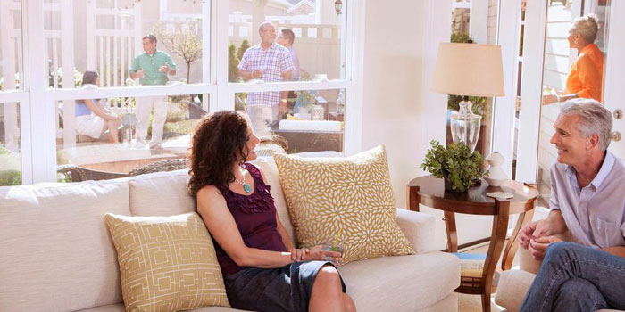 A view of a small get-together. Foregrounds is of a woman sitting on a couch, in front of a window, talking to a man in an armchair. Outside the window, four people have gathered.
