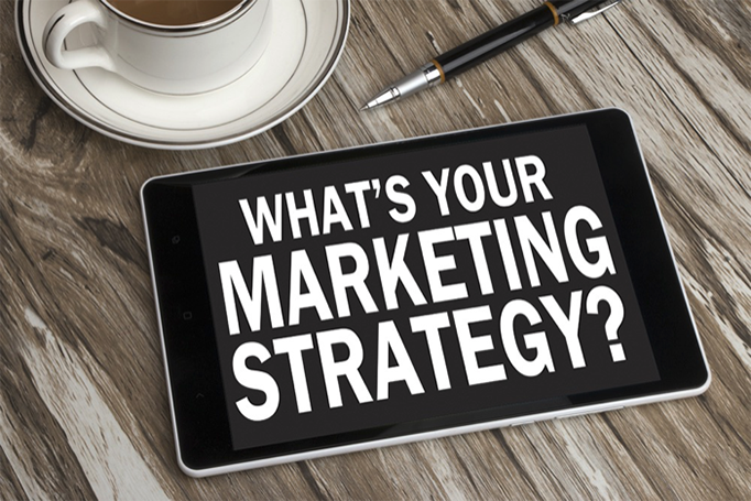 A tablet reading WHAT'S YOUR MARKETING STRATEGY? sitting on a wooden table near a pen and cup of coffee